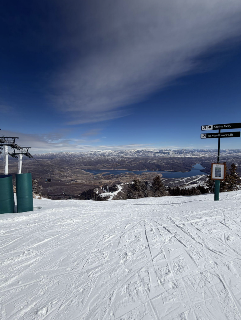 Snow-covered ski slope with a chairlift on the left, directional signs on the right, and mountains and a lake in the background under a partly cloudy sky—perfect for any ski and snowboard club outing.